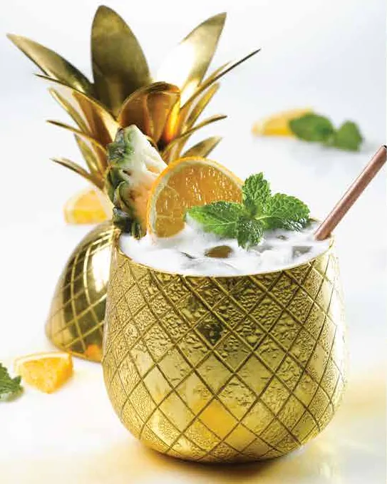 A gold pineapple-shaped cup filled with a creamy cocktail, garnished with orange slices and mint