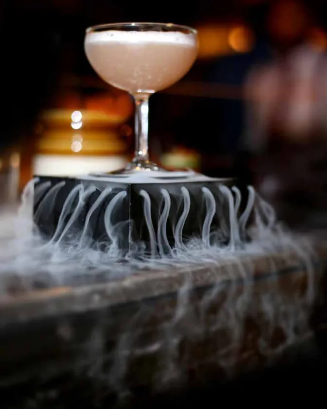A pale coupe cocktail on a black stand wreathed in dry-ice smoke, in a dim bar
