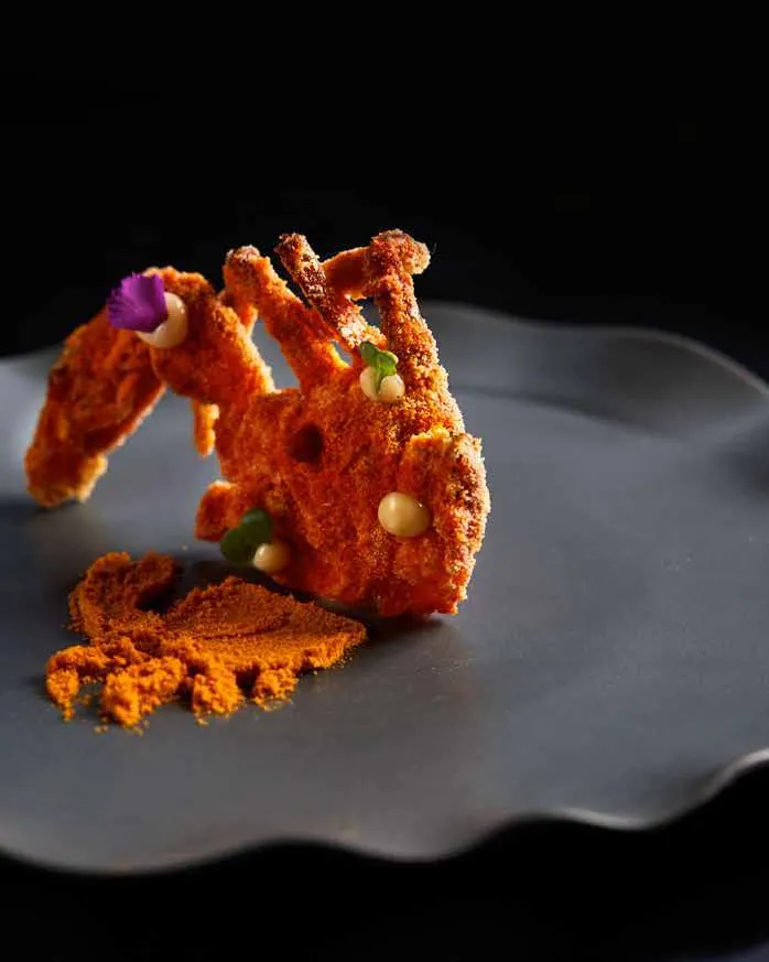 Podi-crusted soft shell crab on a grey wavy plate with a smear of orange spice powder, against a black background