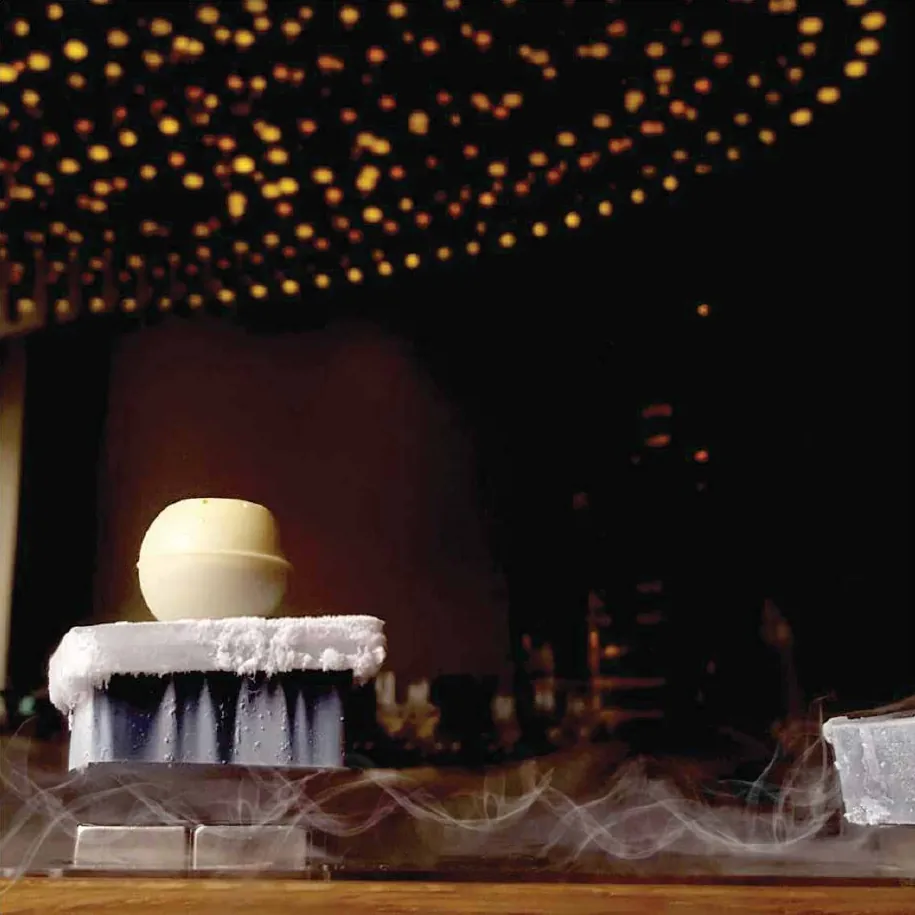 A white dessert sphere resting on a frosted ice block with dry-ice mist, lit by warm bokeh lights in a dark room