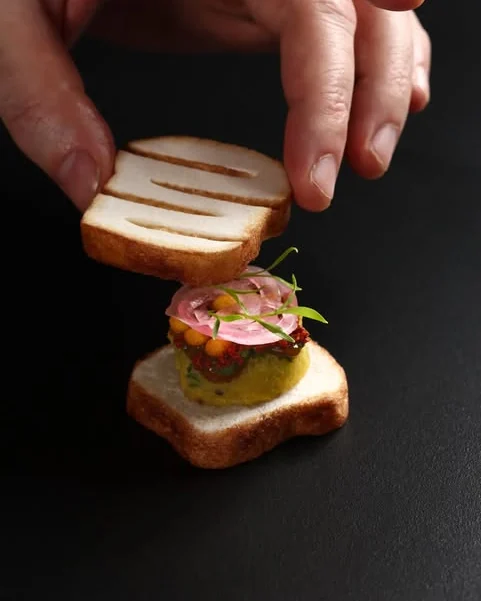 A hand placing a grill-marked onion-water meringue “toast” over aloo bonda topped with pickled onion, chutney and micro-greens, on a black surface
