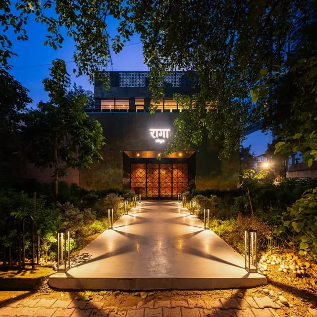 Raga by Gaggan at dusk: a lit stone pathway through a garden leads to the illuminated राग sign and copper lattice doors