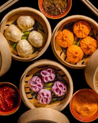 Bamboo steamers of white, orange and pink flower-shaped momos with dipping sauces